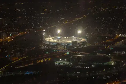 A cricket stadium under floodlights, seen from above at night