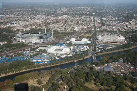 An aerial view of Melbourne Park beside the Yarra River, with the tennis arenas, blue courts and the cricket ground beyond