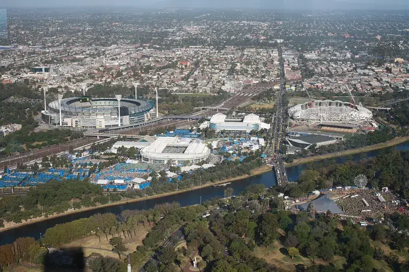 An aerial view of Melbourne Park beside the Yarra River, with the tennis arenas, blue courts and the cricket ground beyond
