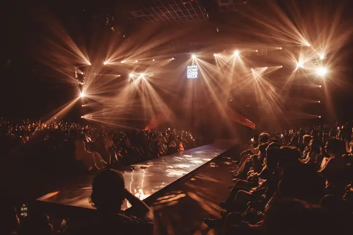 A fashion-show runway under dramatic stage lights, with the audience in shadow